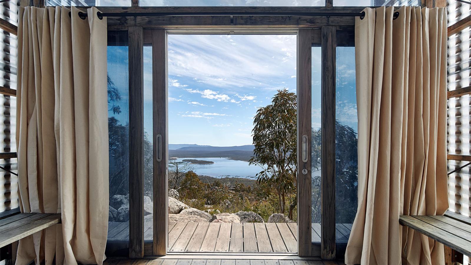 A set of glass double doors with beige curtains open to a deck, overlooking a scenic view of a lake surrounded by the majestic Gariwerd hills and trees under a blue sky with wispy clouds. The foreground features rocky terrain leading to this serene landscape, reminiscent of the Grampians Peaks Trail.