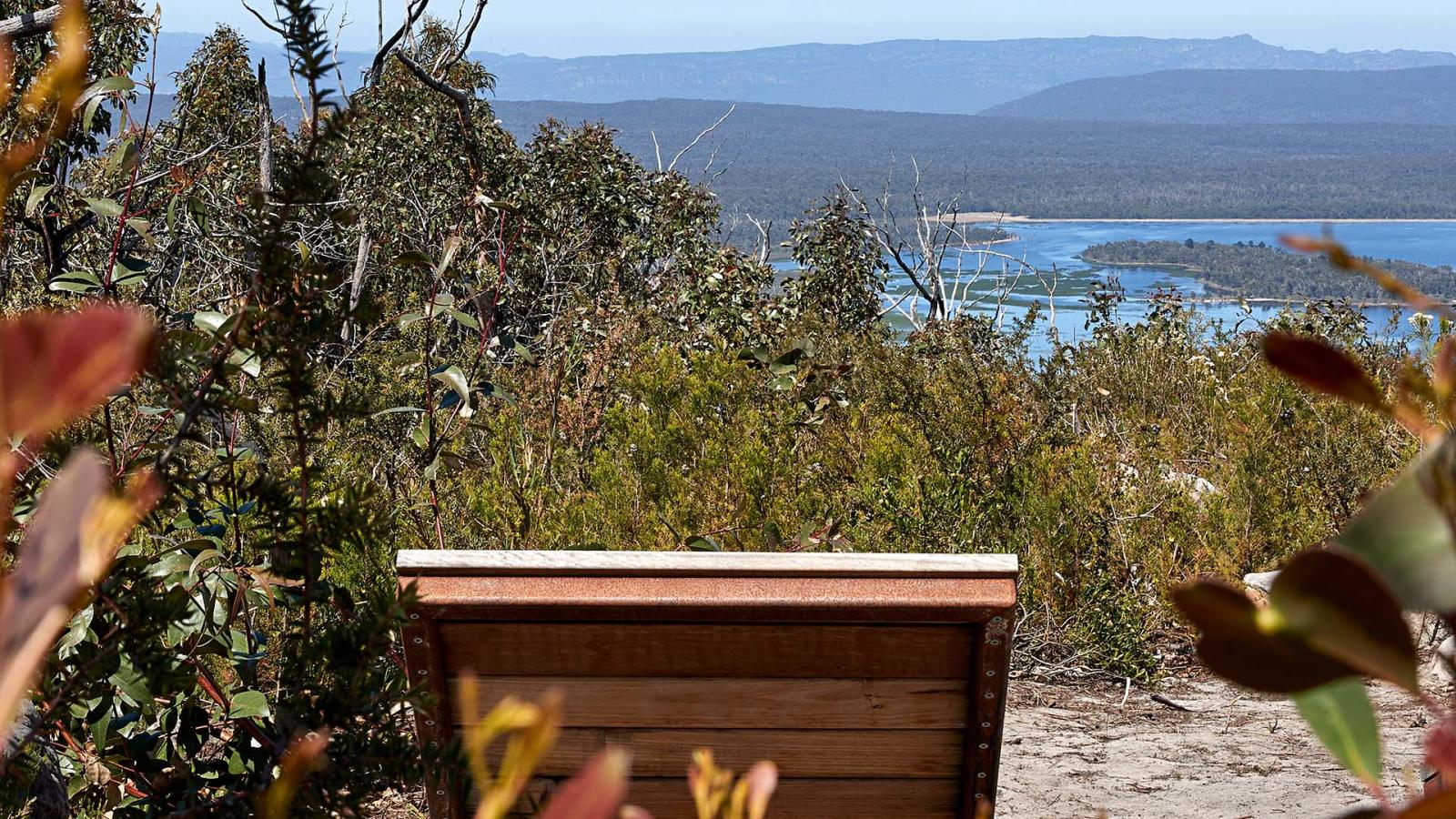 A wooden bench at a scenic overlook, surrounded by foliage, offers a panoramic view of a vast landscape. In the distance, a serene body of water is nestled amid the rolling hills and mountains of Gariwerd under a clear blue sky.