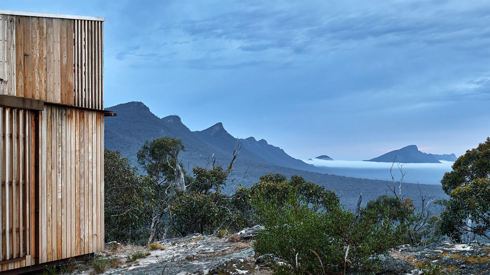 A modern wooden building with slatted exterior walls is set against the mountainous landscape of Gariwerd. The sky is overcast, and a layer of clouds stretches between the peaks of the Grampians Peaks Trail. Trees and shrubs fill the foreground, adding natural elements to the scene.