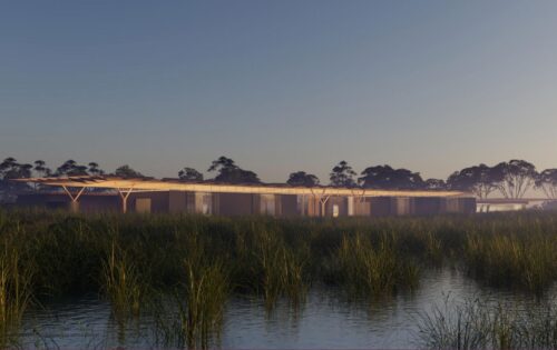 A modern, low-lying building with a flat roof and large glass windows is set against the serene Hobsons Bay Wetlands at sunset. The Centre is surrounded by tall grasses and water, with trees in the background under a dusky sky.