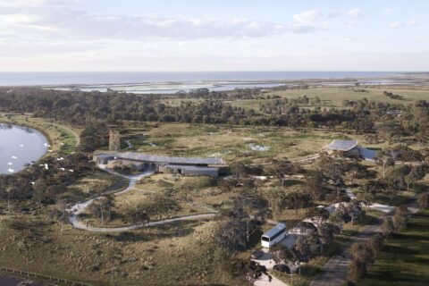 Aerial view of the Hobsons Bay Wetlands Centre featuring a modern building with a green roof, surrounded by lush vegetation and wetlands. A winding pathway weaves through the landscape, with water bodies and open fields extending to the horizon.