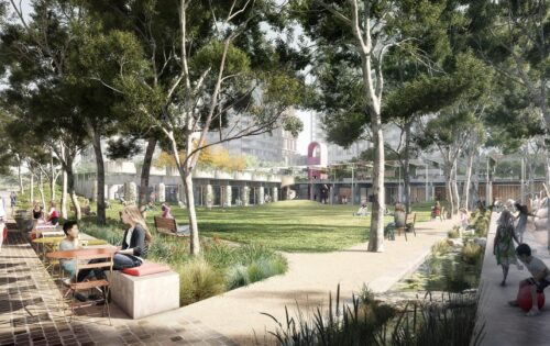 A lively park features people enjoying outdoor activities. Some sit at tables near the walkway, while others relax on benches. Children play in the green space and on a playground. Trees provide shade, and buildings are visible in the background of Central Park.