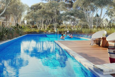 A serene outdoor pool in the heart of Leichhardt, surrounded by lush greenery and trees. Two lounge chairs and a table with a basket are on the wooden deck. People are enjoying the pool; one person is floating on a chair and another is holding a beach ball. Towels are neatly folded nearby.