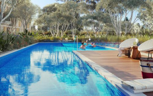 A serene outdoor pool in the heart of Leichhardt, surrounded by lush greenery and trees. Two lounge chairs and a table with a basket are on the wooden deck. People are enjoying the pool; one person is floating on a chair and another is holding a beach ball. Towels are neatly folded nearby.