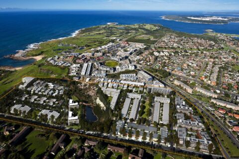 Aerial view of an urban coastal area featuring a community with houses, roads, and green spaces. A large section marked by a yellow outline at Little Bay Cove contains buildings arranged in a grid pattern. The coastline and ocean are visible in the background.