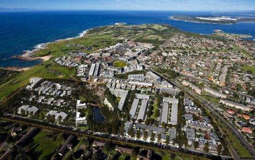 Aerial view of an urban coastal area featuring a community with houses, roads, and green spaces. A large section marked by a yellow outline at Little Bay Cove contains buildings arranged in a grid pattern. The coastline and ocean are visible in the background.