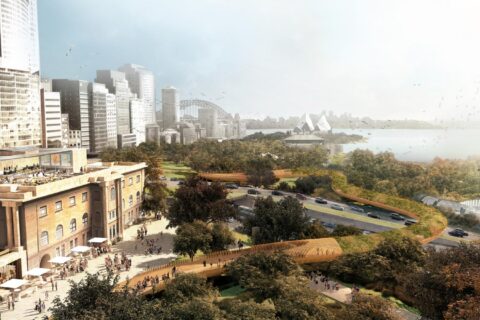 An urban park with trees and pathways overlooks a city skyline with tall buildings. A bridge and a renowned opera house are visible in the background near a body of water. As part of the precinct’s revitalisation, people walk along Macquarie St's paths, while some sit at an outdoor café.