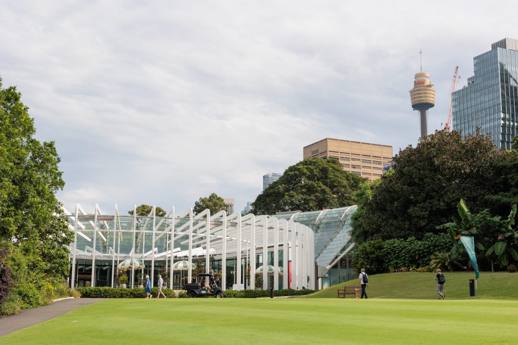 A modern glass building with white supports stands elegantly in a lush garden at the Royal Botanic Gardens. People stroll nearby, and a golf cart glides along the path. Towering city skyscrapers, including a needle-like tower, rise majestically in the background under a cloudy sky.