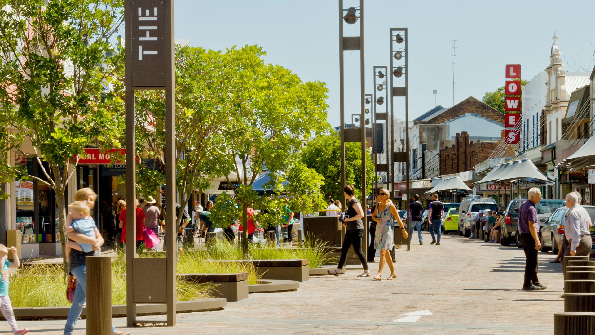 A bustling outdoor shopping street in Maitland with people walking, some holding children. Tall, stylish light posts and greenery line the street alongside various shops and restaurants. The sky is clear, making it a perfect sunny day by the levee.
