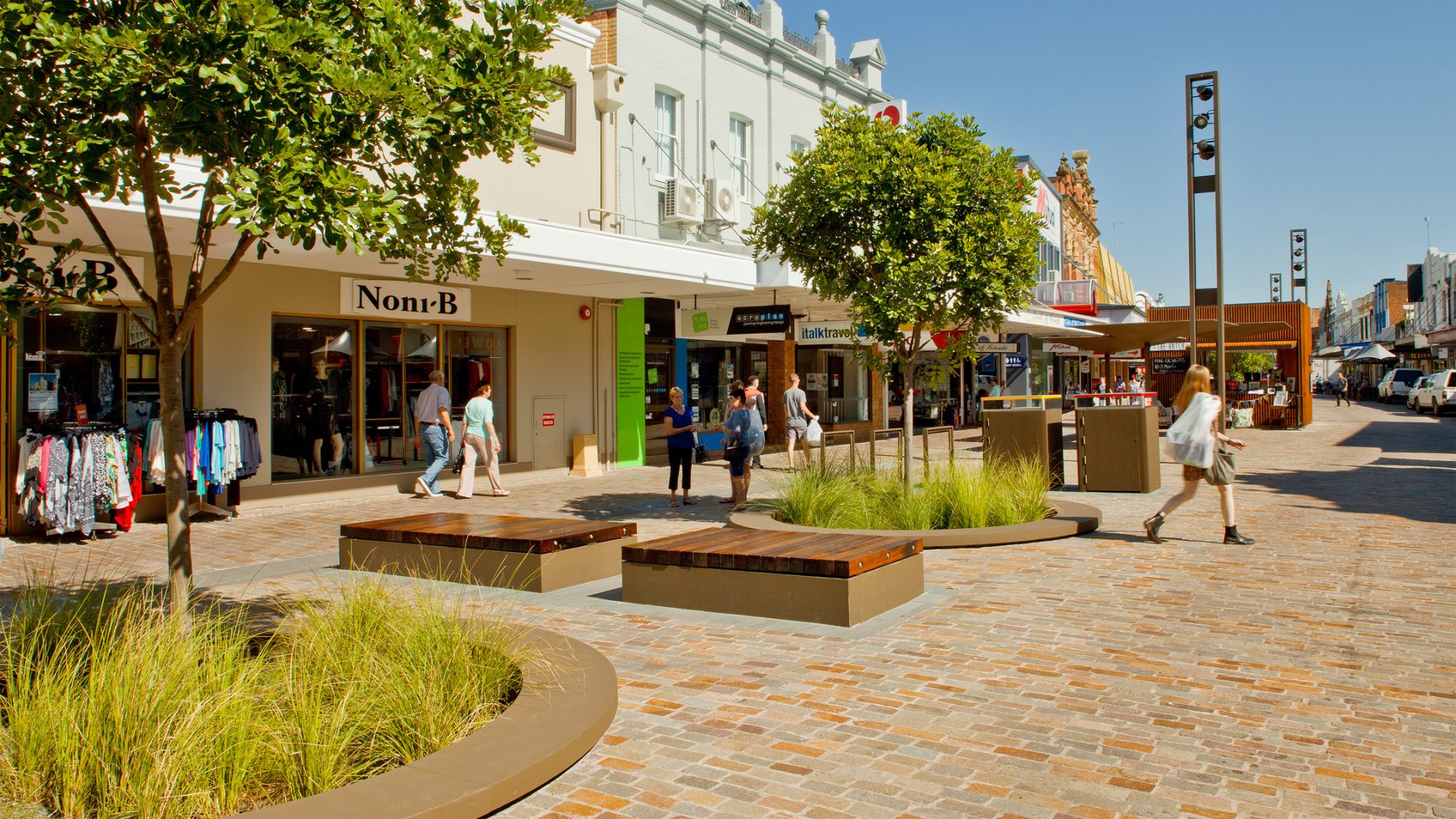 A lively pedestrian shopping street in Maitland features trees and modern benches surrounded by greenery. Stores, including a clothing store named Noni B, line both sides of the Levee. Several people are walking, browsing, and relaxing in the pleasant, sunny environment.