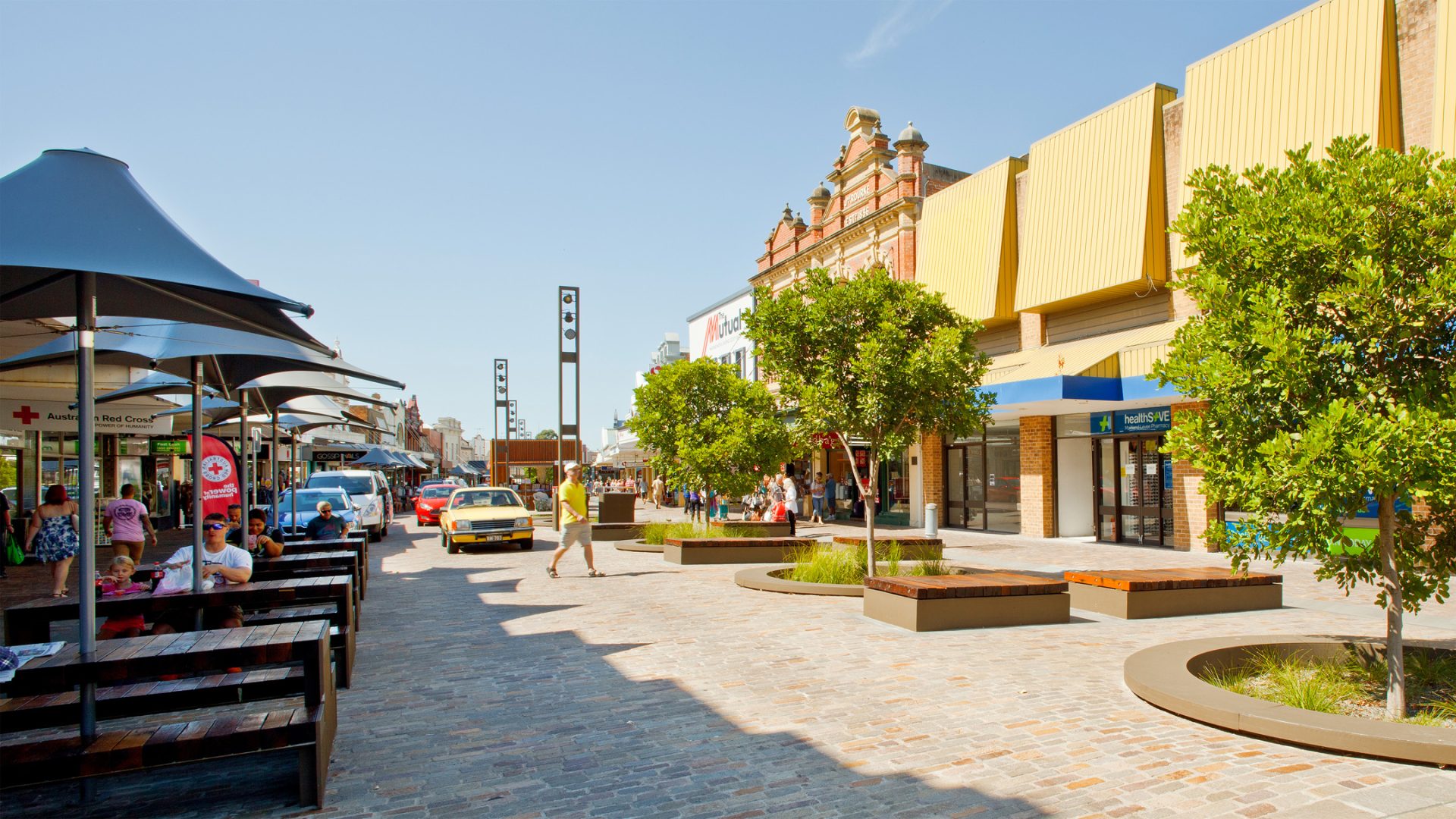 A lively outdoor shopping street in Maitland features modern storefronts, trees in circular planters, and patio umbrellas shading tables with people dining. A yellow car is parked by the levee, while pedestrians stroll along the brick-paved walkway under a clear blue sky.