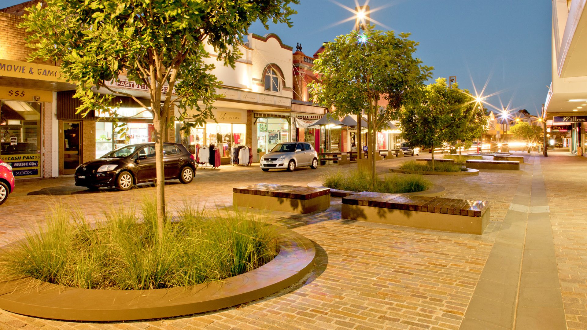 A well-lit, cobblestone street in Maitland, lined with trees and benches at dusk. Retail shops with large windows displaying clothing are on the left, and cars are parked along the side. A few pedestrians are walking along the levee, creating a calm, inviting atmosphere.