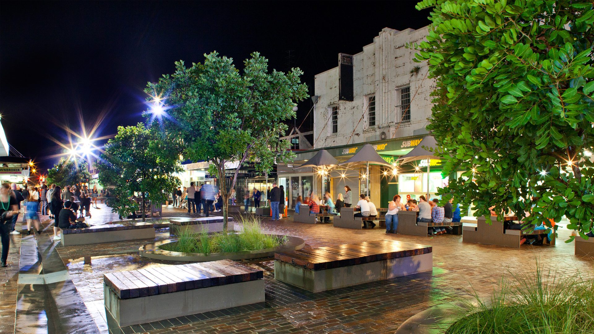 A bustling outdoor plaza at night, with people dining at brightly lit restaurants and walking along the pathway. Benches and trees are scattered around the area, with lights twinkling from the restaurant on the right side. The lively scene could easily be from a charming spot in Maitland.
