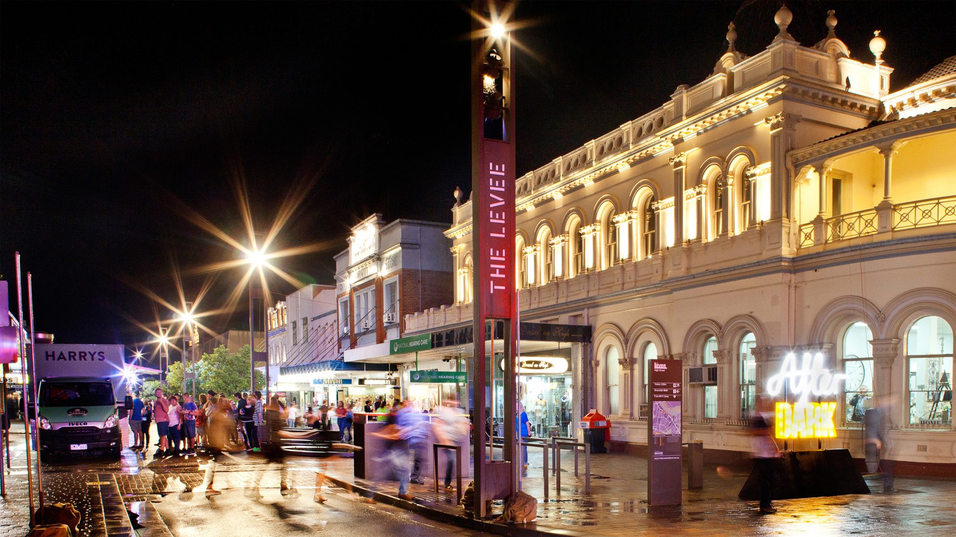 A lively, bustling street scene at night, lined with illuminated buildings and shops. Crowds of people are seen walking and gathering. A prominent building on the right, reminiscent of Maitland's classical architecture, stands tall. Near the center, a lit sign reads "Hello DARKNESS.