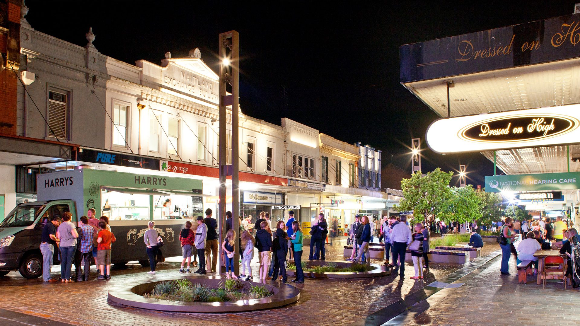 A vibrant street scene at night with people gathered near food trucks and outdoor seating areas on Maitland Levee. The street is illuminated with bright lights and various shops and cafes are visible. A sign for "Dressed on High" hangs prominently in the foreground.