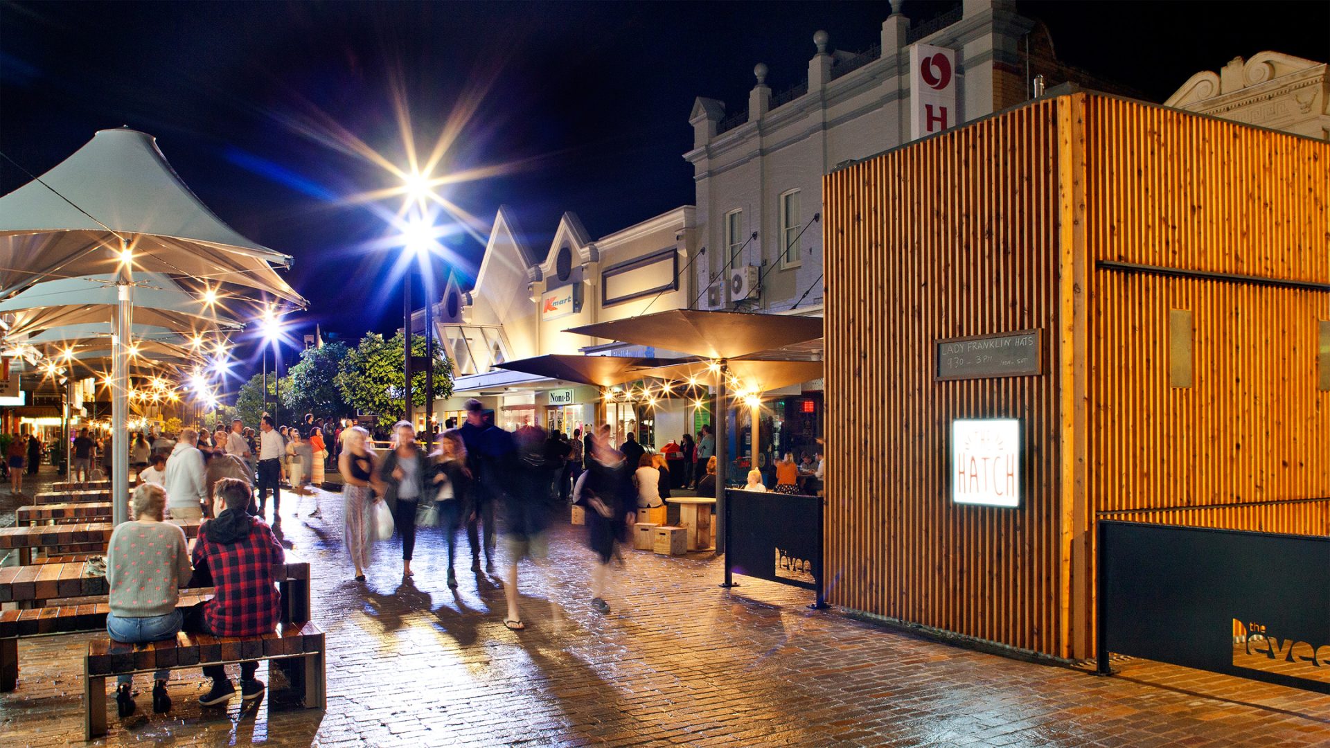 A bustling Maitland street at night with numerous people walking and sitting on benches. The street is lined with shops and restaurants, some with outdoor seating. Bright lights and lanterns illuminate the area, giving it a lively atmosphere. A wooden structure is prominently visible by the levee.
