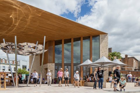 A modern building with a wooden overhang and large glass windows stands under a partly cloudy sky in Maitland. People gather in the open area, some shaded by large geometric sculptures and umbrellas. The scene showcases a vibrant, community-oriented space enhanced by the nearby Levee and contemporary architecture.