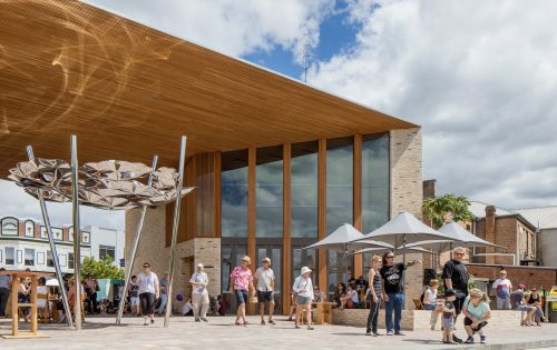 A modern building with a wooden overhang and large glass windows stands under a partly cloudy sky in Maitland. People gather in the open area, some shaded by large geometric sculptures and umbrellas. The scene showcases a vibrant, community-oriented space enhanced by the nearby Levee and contemporary architecture.