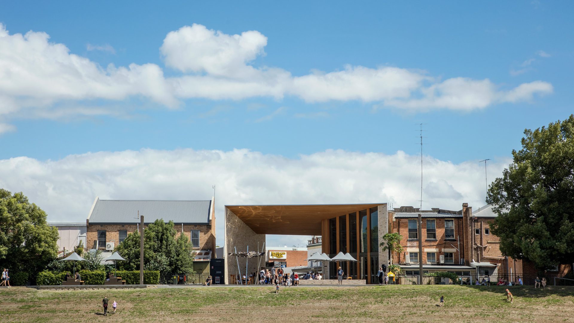 A modern building with a large, open wooden structure and an overhanging roof stands in an urban area. Surrounding it are older brick buildings. People are visible in a grassy area near the levee in the foreground under a blue sky with scattered clouds.