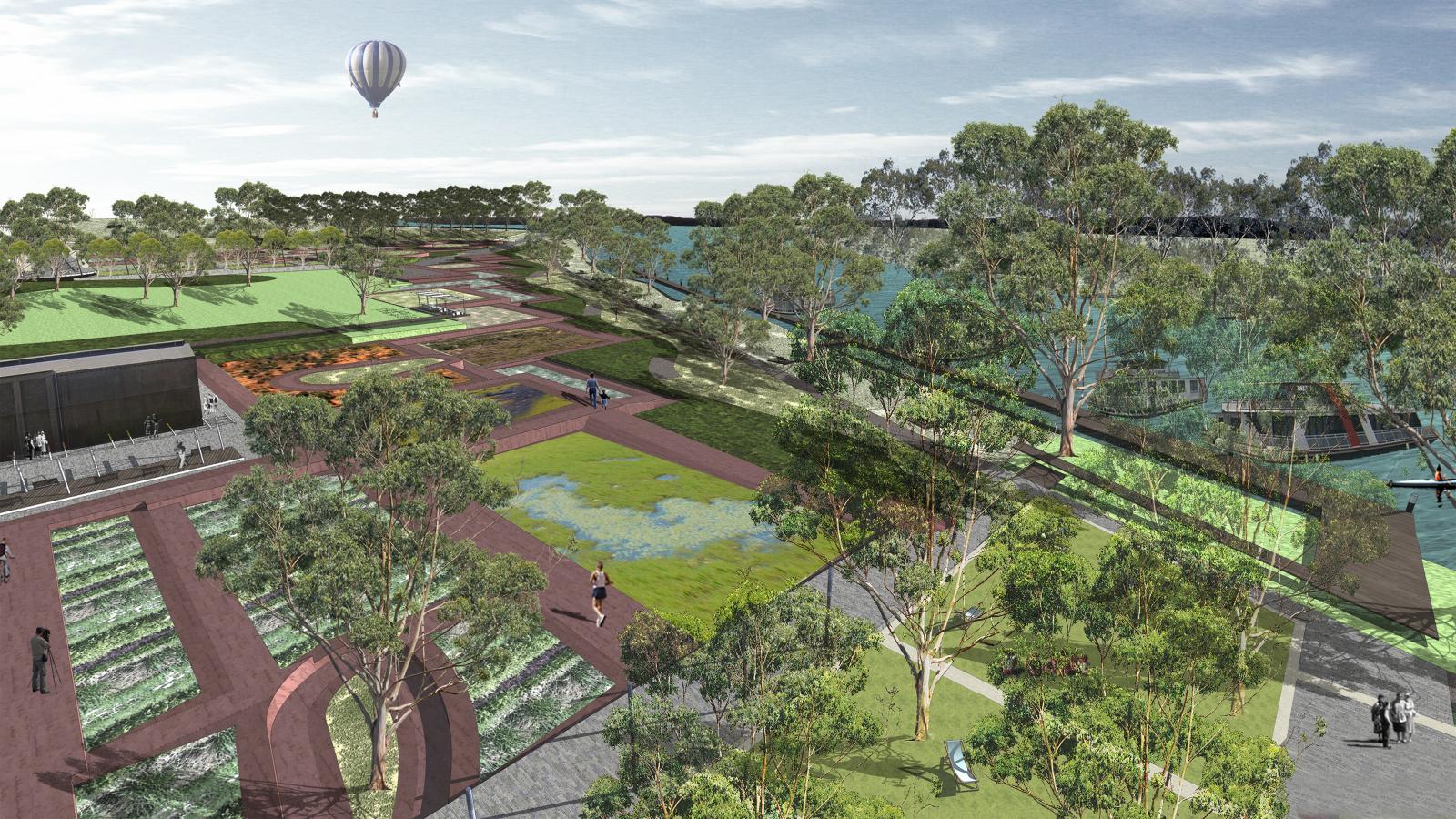 A scenic park with gardens, lawns, and walking paths beside the Mildura riverfront. People are enjoying the space, some sitting and others walking. Trees line the park, and a hot air balloon floats in the sky above. A few boats are seen on the river.