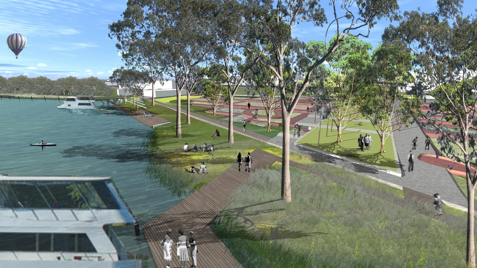 An urban riverside park in Mildura features a walking path along the water, trees, grassy areas, and people enjoying various activities like walking, sitting, and kayaking. A hot air balloon is visible in the sky, and a modern structure is partially seen in the foreground of this picturesque riverfront setting.