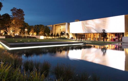 A modern building with large illuminated panels is situated near a serene reflecting pool at dusk. The structure, reminiscent of the elegant design of the NGA, is surrounded by trees and a well-lit Australian garden, adding to the tranquil ambiance. The clear sky and calm water create a peaceful scene.