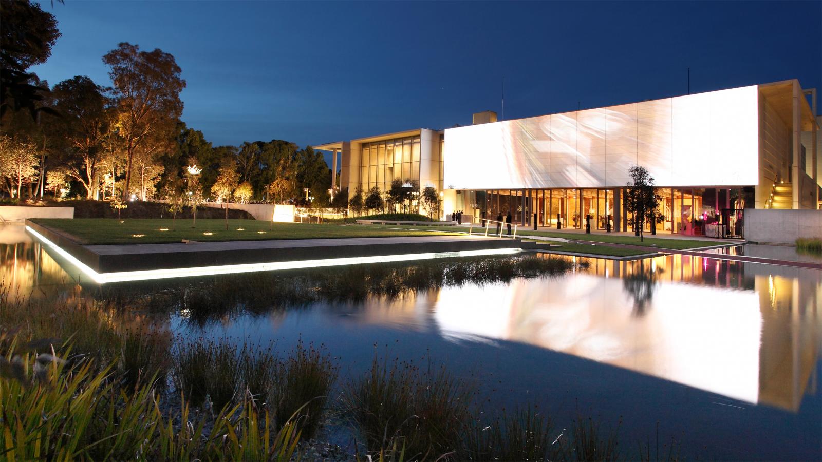 A modern building with large illuminated panels is situated near a serene reflecting pool at dusk. The structure, reminiscent of the elegant design of the NGA, is surrounded by trees and a well-lit Australian garden, adding to the tranquil ambiance. The clear sky and calm water create a peaceful scene.