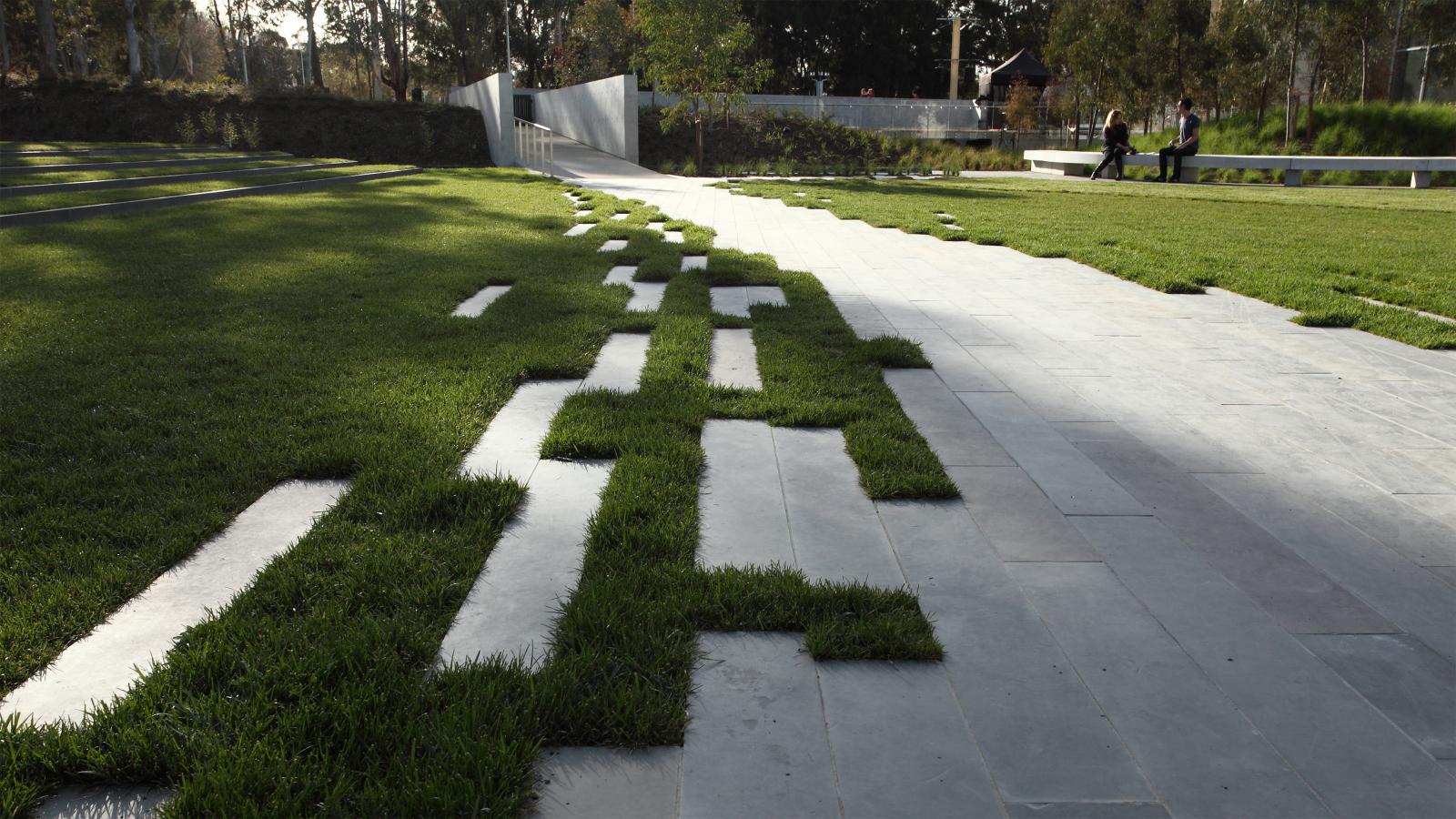 A contemporary park featuring a unique design of interlocking grass and concrete paths, reminiscent of the Australian Garden at NGA. In the background, two people are talking on a white bench. The park is surrounded by trees and greenery with an open metal gate visible in the distance.