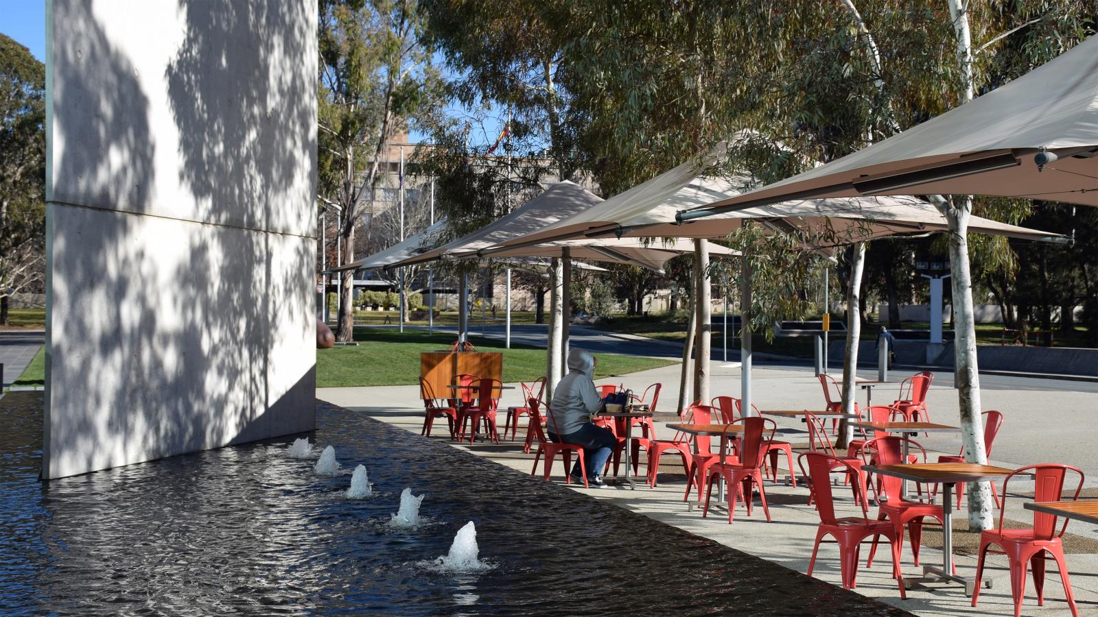 Outdoor café with red chairs and tables, shaded by large umbrellas. A person in a gray hoodie sits alone at a table. The area is adjacent to a fountain with several water jets and surrounded by trees from the Australian Garden at the NGA. Some buildings are visible in the background.
