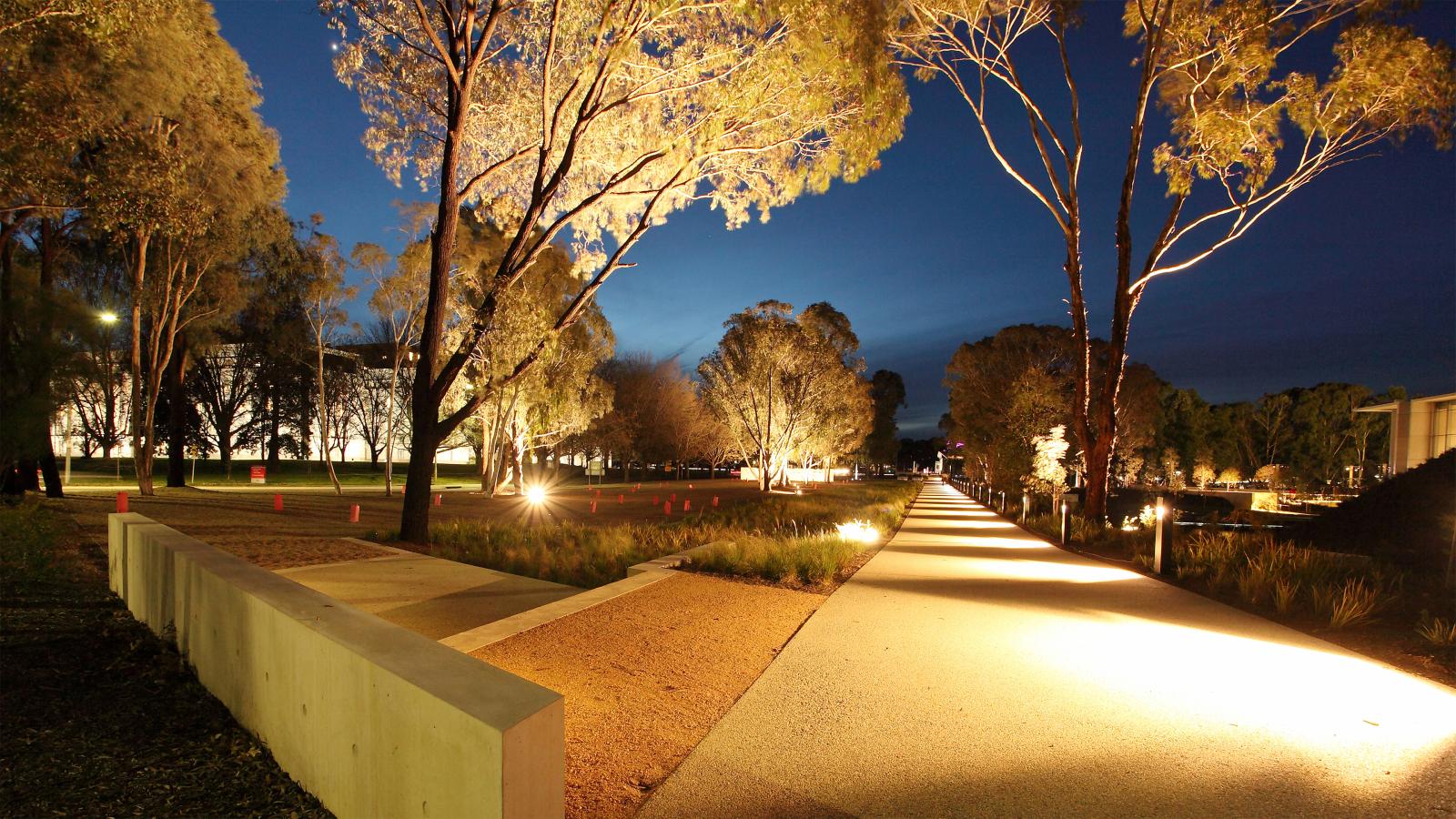 A well-lit pathway through an Australian park at twilight, with tall trees accentuated by the soft glow of surrounding lights. The sky is deep blue, indicating the transition from day to night. The path leads into the distance, flanked by lush garden vegetation.