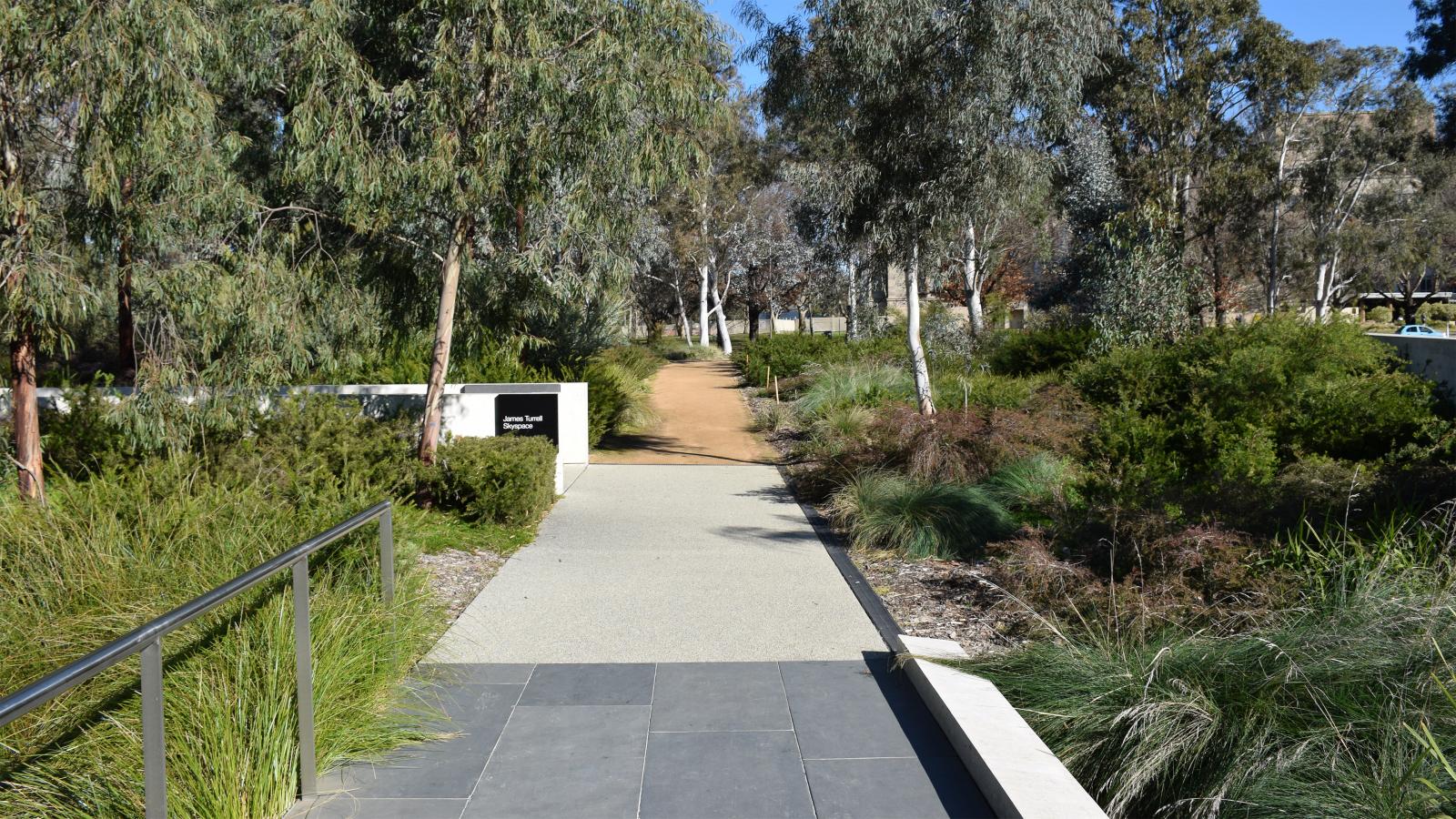A paved pathway extends into a lush garden filled with various green shrubs and trees. A small sign on the left reads "Main Garden Path." The path is partially shaded by tall trees and bordered by railings and low vegetation. In the background, a dirt path leads further into the Australian Garden at NGA.