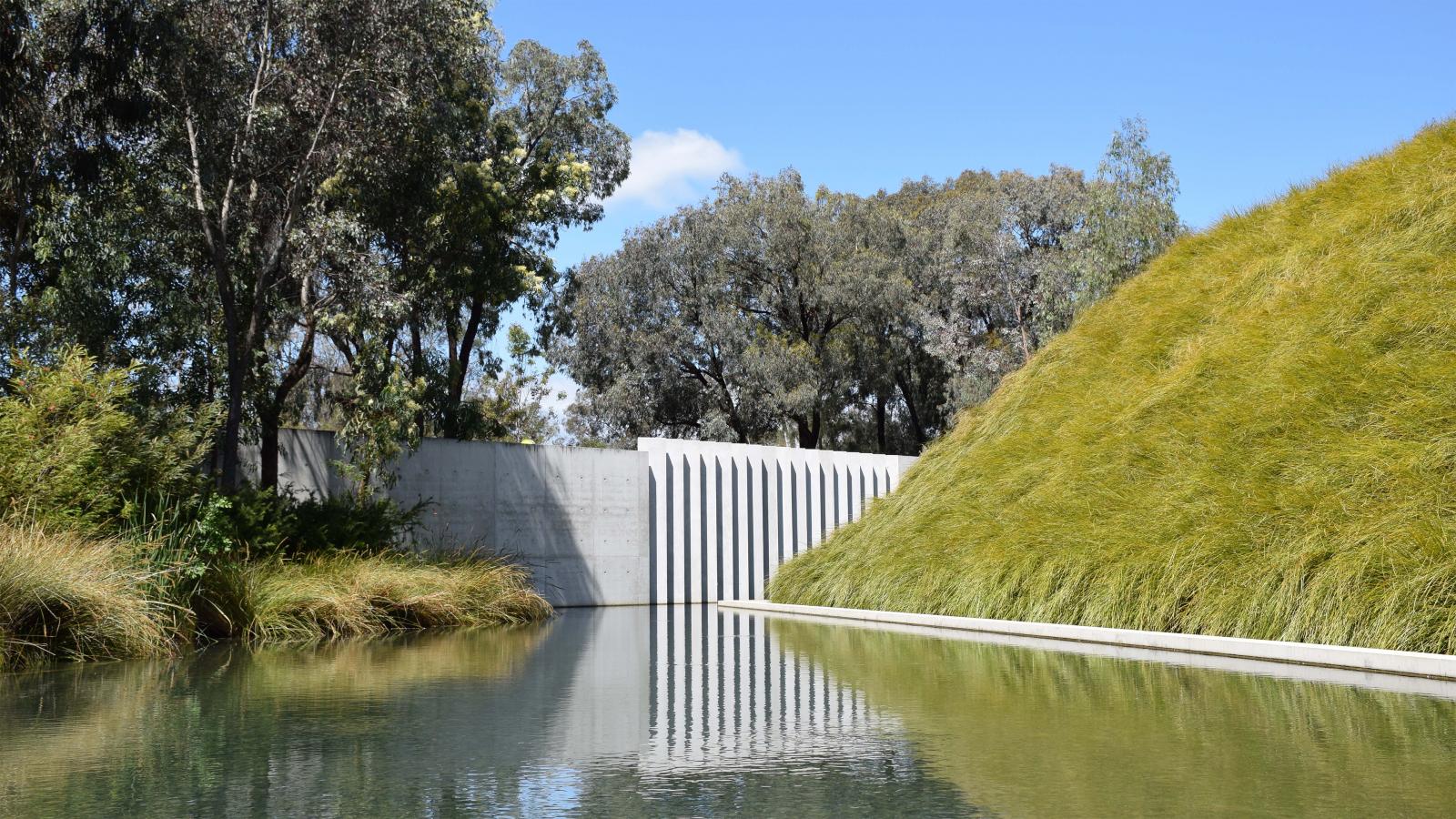 A serene landscape featuring a reflective pond bordered by tall grasses and trees in the Australian Garden. A modern, white, geometric wall with vertical lines stands at the edge of the pond, creating a striking contrast with the natural greenery and blue sky in the background.
