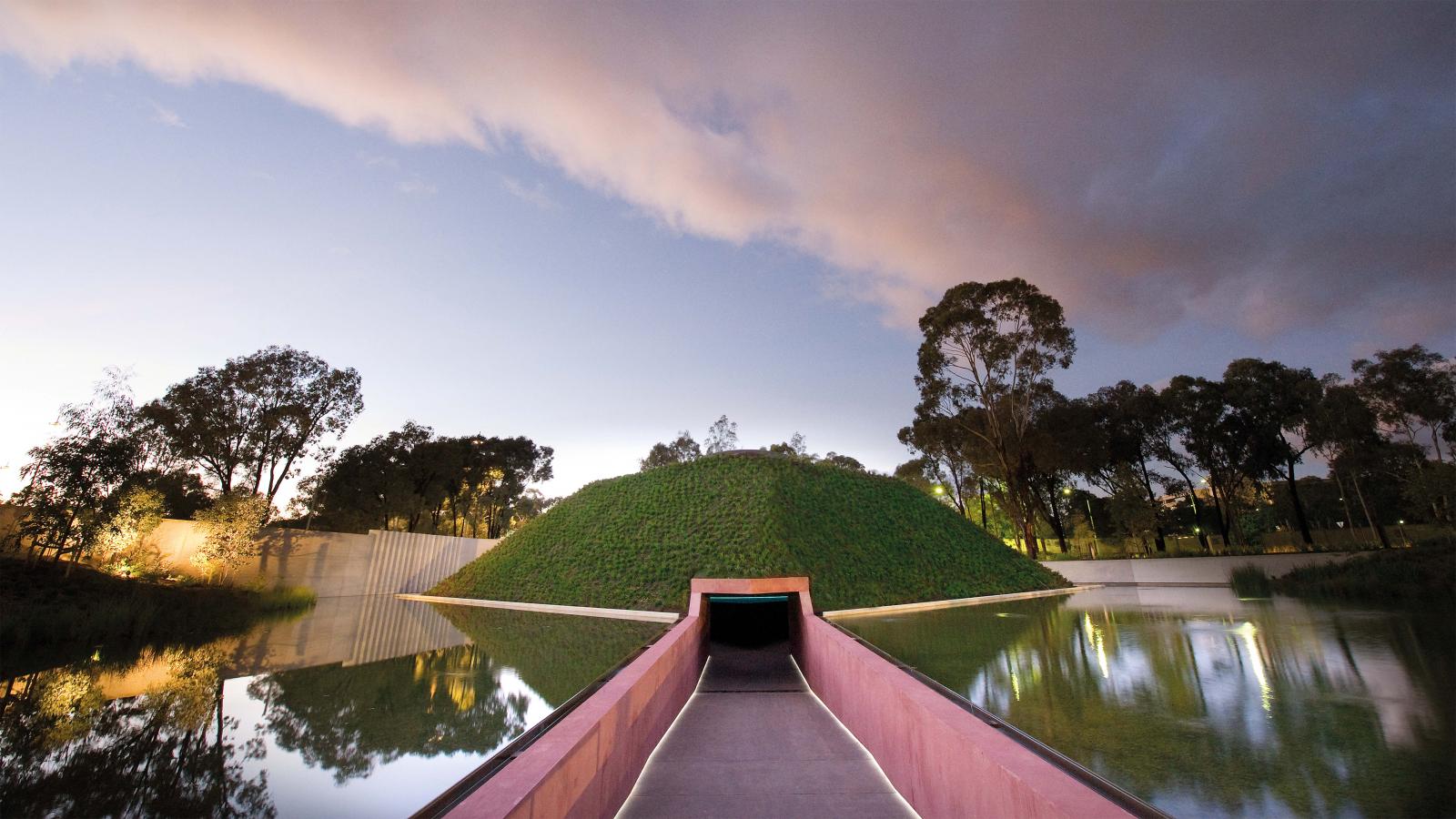 A view of the Skyspace installation at the National Gallery of Australia, featuring a green, grassy mound surrounded by water in the Australian Garden. The sky is partly cloudy and the scene is reflected in the still water, creating a serene and symmetrical composition at NGA.