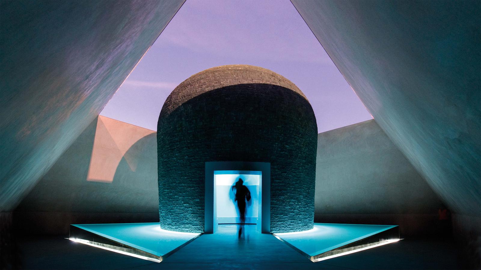 A person walks through a futuristic, dimly lit triangular hallway with a dome-shaped structure in the center, illuminated by blue light. The geometric space features sharp angles, creating a surreal, otherworldly atmosphere reminiscent of an exhibit at the NGA's Australian Garden.