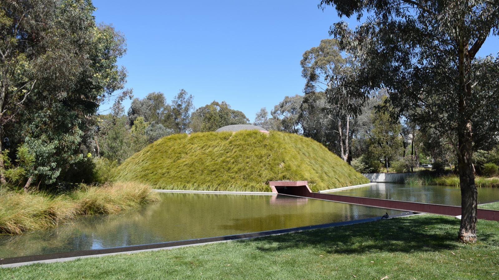 A lush, green grassy mound surrounded by a tranquil pond on a sunny day. A red bridge extends from the mound over the water in this picturesque Australian Garden. Trees and vegetation border the scene, adding a natural ambiance under a clear blue sky at NGA.