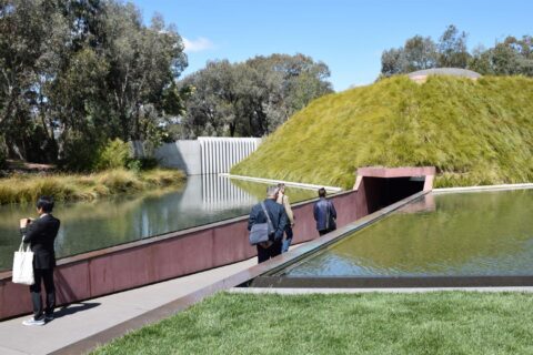 Three people walk towards a modern earth-sheltered building with a grass-covered roof in the Australian Garden, surrounded by water and greenery. One person is taking a photo while others approach the building's entrance. Trees and a blue sky can be seen in the background.