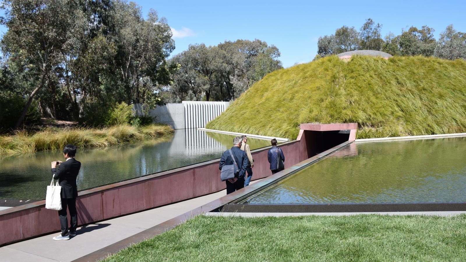 Three people walk towards a modern earth-sheltered building with a grass-covered roof in the Australian Garden, surrounded by water and greenery. One person is taking a photo while others approach the building's entrance. Trees and a blue sky can be seen in the background.