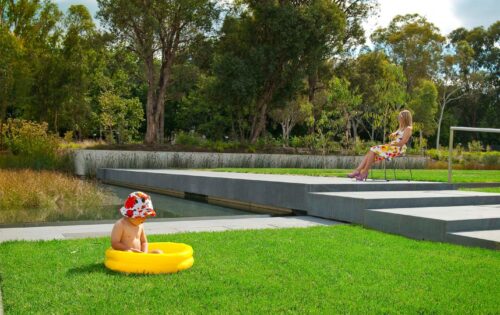 A child wearing a floral hat sits in a small yellow inflatable pool on a grassy area within the Australian Garden near a water feature. A woman in a floral dress sits on a chair on a concrete platform overlooking the scene, with trees and greenery in the background, reminiscent of an NGA exhibit.