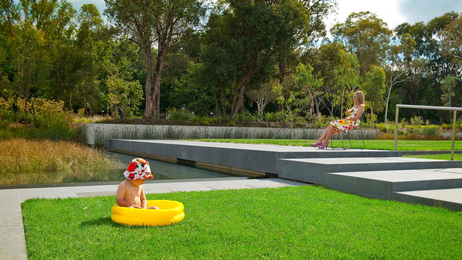 A child wearing a floral hat sits in a small yellow inflatable pool on a grassy area within the Australian Garden near a water feature. A woman in a floral dress sits on a chair on a concrete platform overlooking the scene, with trees and greenery in the background, reminiscent of an NGA exhibit.