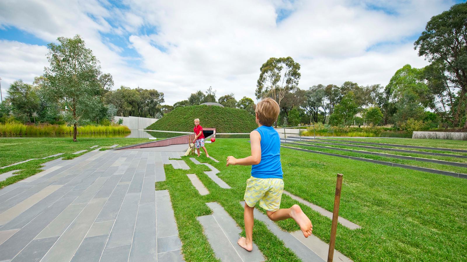Two children play joyfully near a sidewalk in the spacious Australian Garden. One child, wearing a red shirt, looks ready to catch or throw a ball, while the other child, in a blue shirt, runs barefoot on the grass. The lush park features manicured lawns, trees, and a large bushy mound near the NGA.