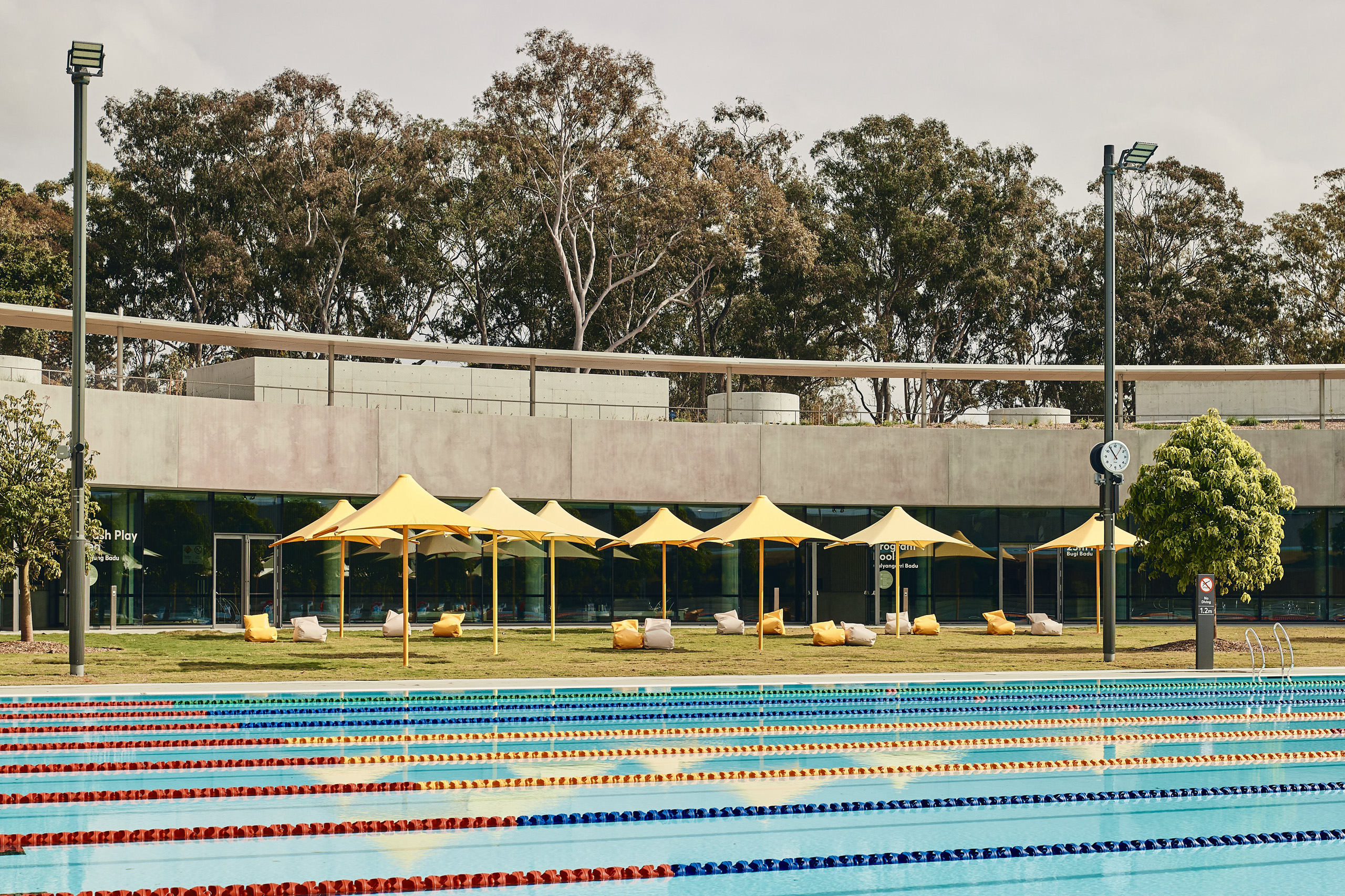 An outdoor swimming pool with colored lane markers in the foreground. Behind the pool at the Parramatta Leisure Centre are several yellow umbrellas and lounge chairs on a lawn. In the background, there is a modern building surrounded by trees. The sky is overcast.