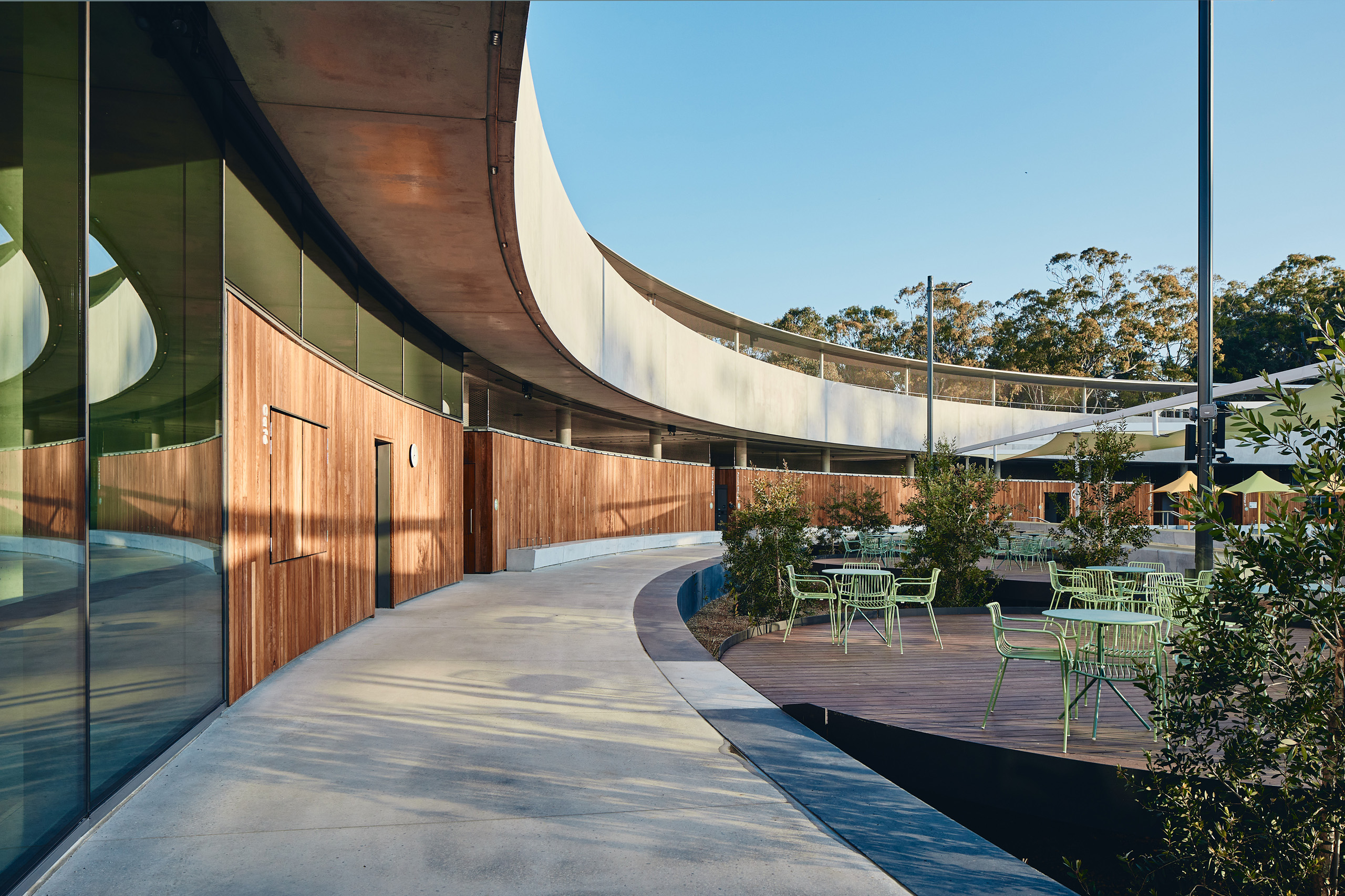 The image shows a modern leisure centre in Parramatta with a curved glass facade and wooden walls. In the foreground, there's an outdoor sitting area with green chairs and tables on a wooden platform surrounded by lush greenery. The sky is clear and blue, creating a serene atmosphere.