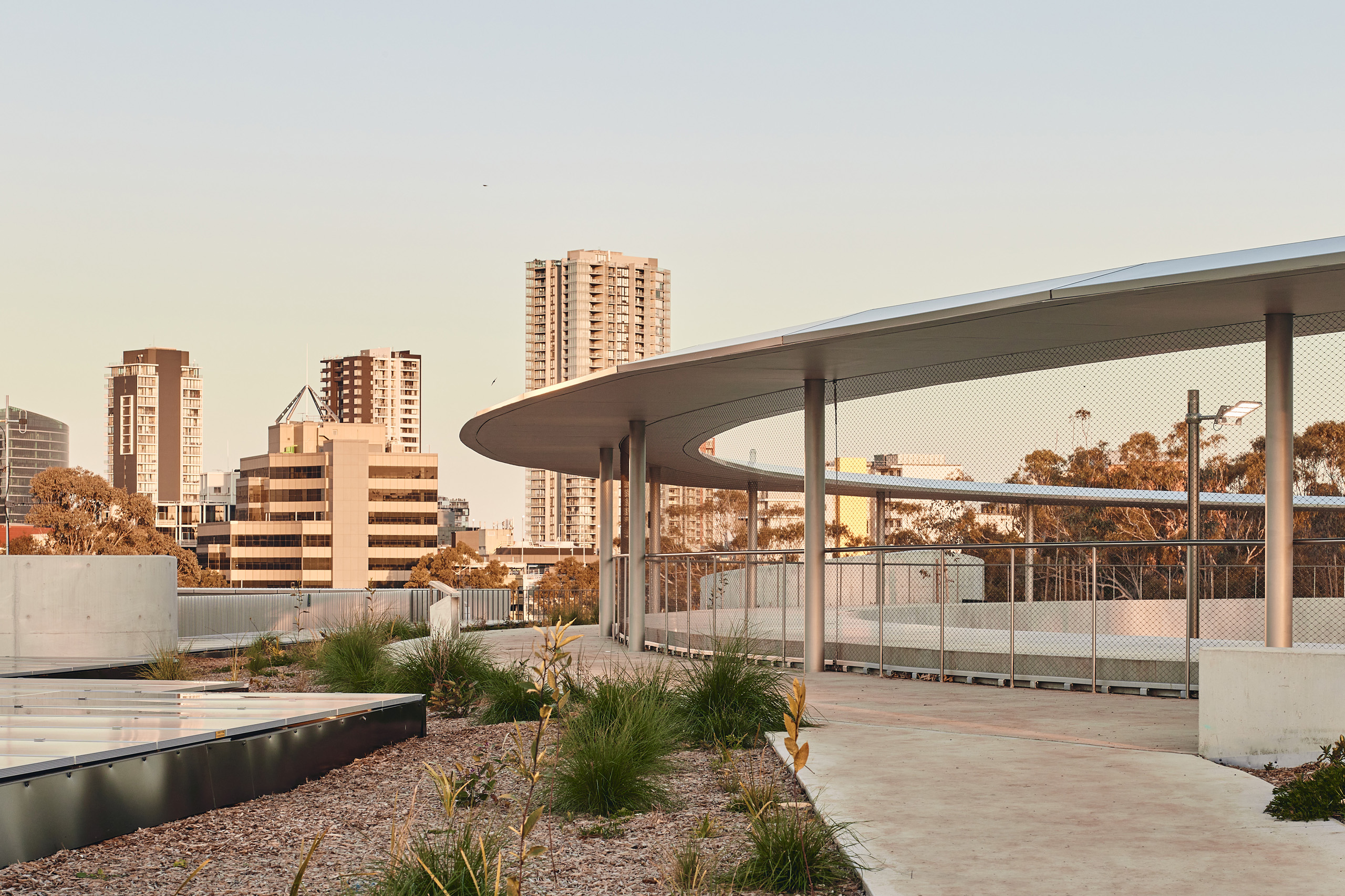 A modern architectural structure with curved, open-air walkways, featuring metal railings and a rooftop garden with sparse vegetation. The backdrop includes tall city buildings and a clear sky, indicating an urban setting. This picturesque scene is part of the Parramatta Aquatic Leisure Centre.