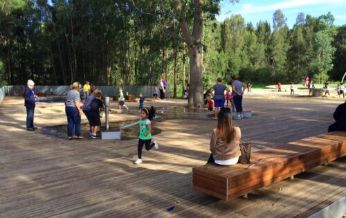 A park scene within a vibrant precinct has people engaged in various activities on a wooden deck. Children are running, playing, and interacting with sandbox features. Adults are standing, walking, and sitting on benches. Trees and greenery surround the area beneath the clear, sunny sky.