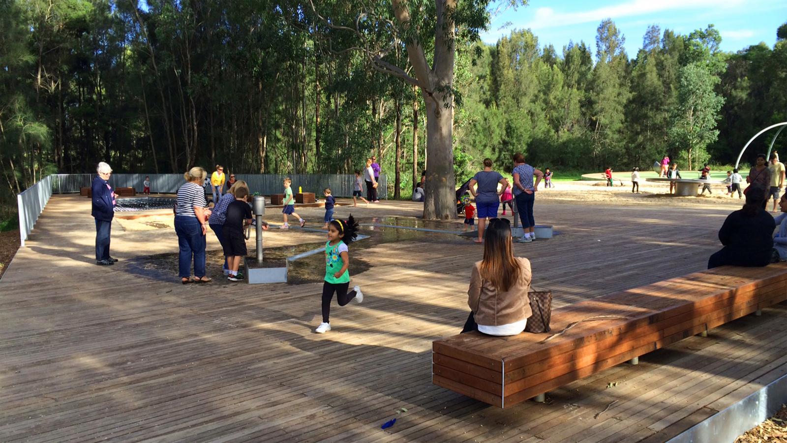 A park scene within a vibrant precinct has people engaged in various activities on a wooden deck. Children are running, playing, and interacting with sandbox features. Adults are standing, walking, and sitting on benches. Trees and greenery surround the area beneath the clear, sunny sky.