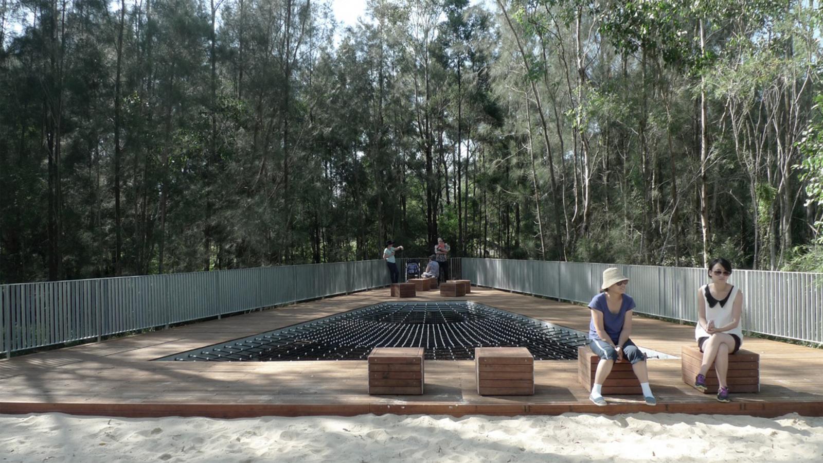 Two people sit on wooden benches on a walkway with a grid of small black cylinders. The platform, reminiscent of a relaxed paddock precinct, is surrounded by trees and more people can be seen standing at the far end. There is sand in the foreground and metal railings along the edges of the walkway.