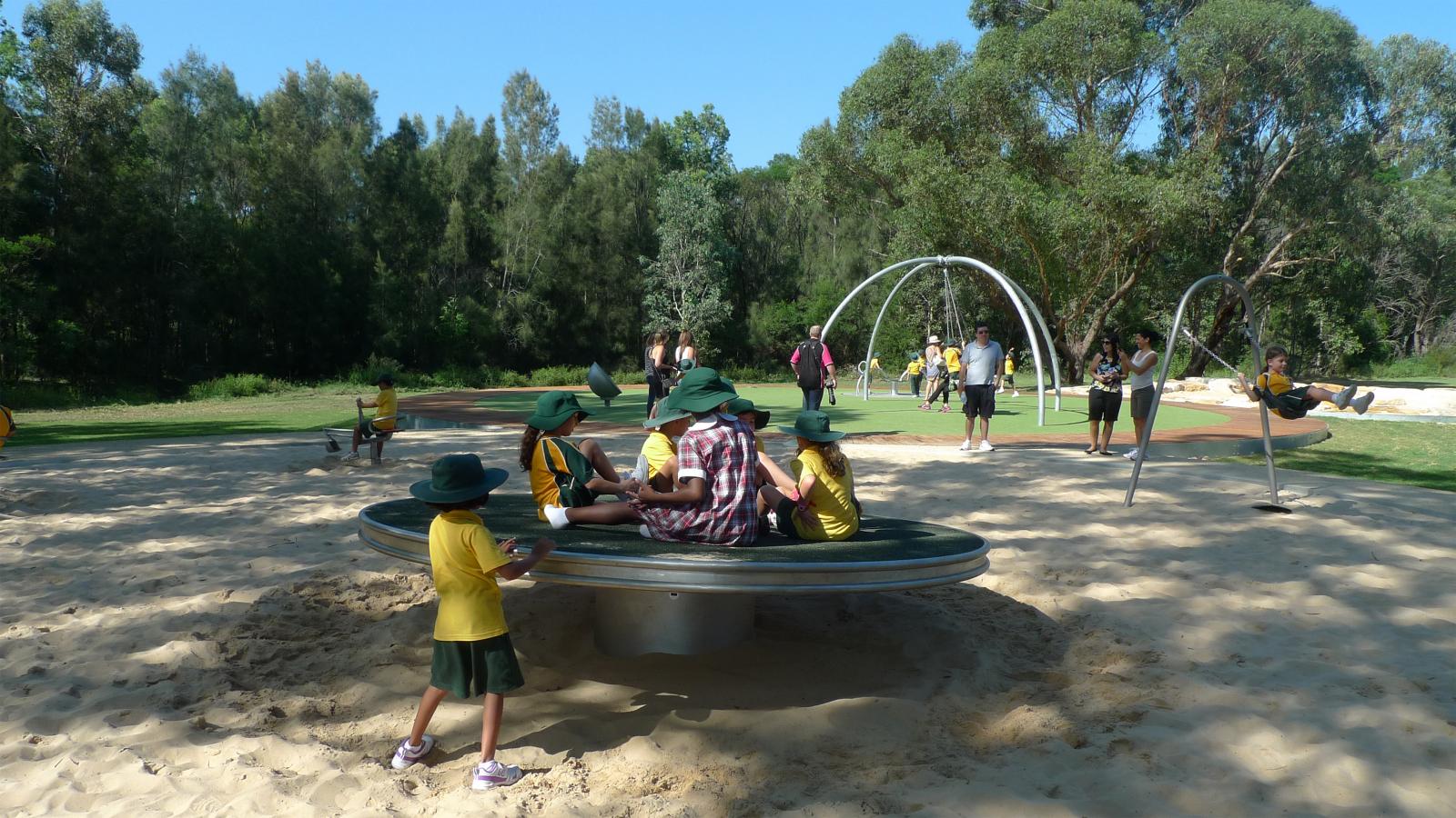 At Paddocks Precinct, a group of children in green hats and uniforms play on a round spinning playground equipment in a sandy area surrounded by trees. Other children and adults are scattered across the playground, enjoying various activities in the background.
