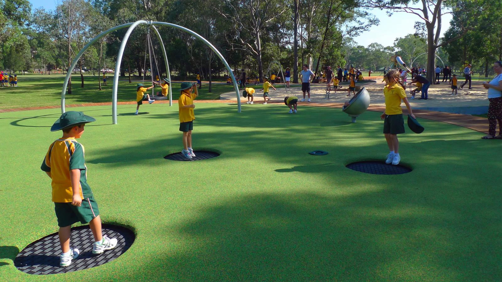 Children dressed in green and yellow school uniforms are playing on a playground with equipment such as a spinning apparatus and circular platforms. The playground, located in a park precinct surrounded by trees and open grassy paddocks, has more people visible in the background.