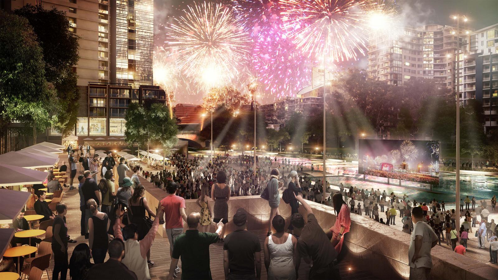 A large crowd gathers in Parramatta near a river to watch a vibrant fireworks display in the night sky. The scene is lively with many people taking photos and enjoying the spectacle. High-rise buildings and illuminated screens can be seen in the background as part of the city's skyline.