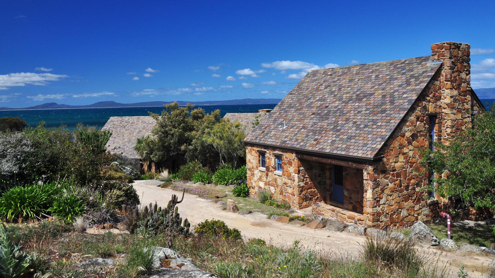 A rustic stone cottage with a shingled roof sits near a dirt path, surrounded by lush greenery and desert plants. This Piermont Retreat overlooks a serene body of water with distant mountains under a clear blue sky, offering an idyllic escape.